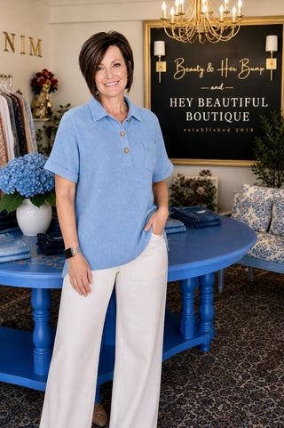 Woman in a blue shirt and white pants standing in a store with a sign that reads 'Hey Beautiful Boutique'.