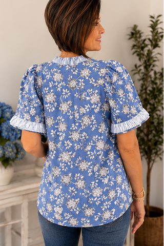 Woman wearing a blue floral blouse with white ruffled trim in an indoor setting.