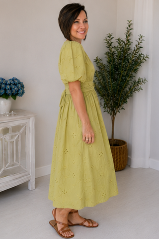 Woman in a yellow dress standing in a room with a white wall, a plant, and a small table with flowers.