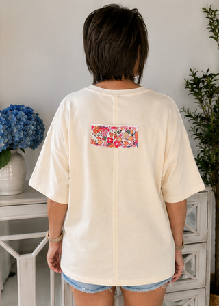 Person wearing a cream-colored t-shirt with a floral pocket design, standing in a room with decorative elements.