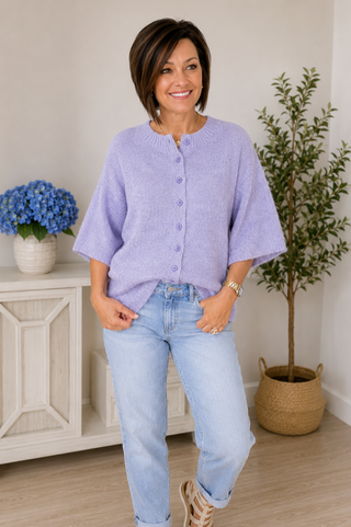 Woman wearing a light purple cardigan and blue jeans standing in a room with a white cabinet and plants.